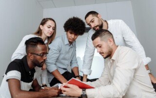 A diverse team of five colleagues gathered around a table reviewing a document together in a bright modern office
